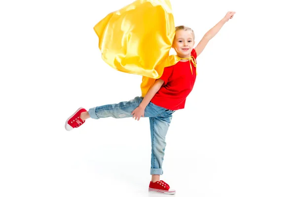 Pequeña superchica sonriente con capa amarilla de pie sobre una pierna y gesto a mano aislado en blanco - foto de stock