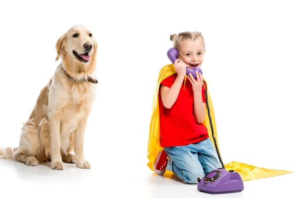 Beige dog with smiling little supergirl talking on phone and wearing yellow cape isolated on white — Stock Photo