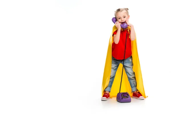 Sorprendido pequeña supergirl usando capa amarilla y hablando por teléfono aislado en blanco — Stock Photo