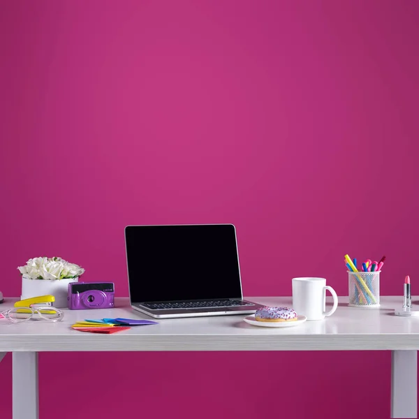 Laptop mit leerem Bildschirm, Donut mit Kaffee, Kamera, Kosmetik und Bürobedarf auf rosa — Stockfoto