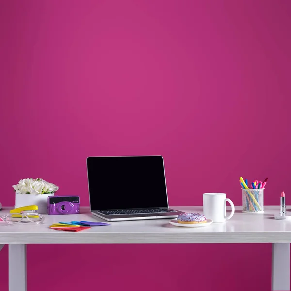 Laptop with blank screen, doughnut with coffee, camera, cosmetics and office supplies on pink — Stock Photo