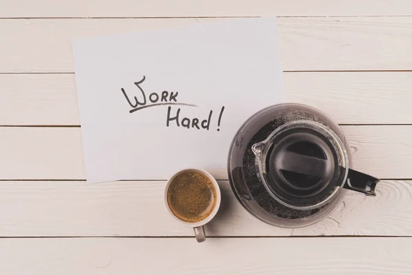 Vista superior de la taza de café, hoja de papel con trabajo de inscripción duro y cafetera en la mesa de madera - foto de stock