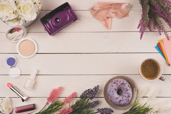 Top view of flowers, camera, cosmetics, cup of coffee and doughnut on wooden table top — Stock Photo