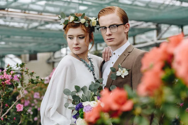 Красивый молодой жених и невеста со свадебным букетом стоят вместе в ботаническом саду — стоковое фото