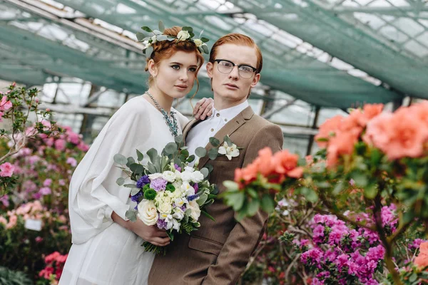 Beautiful elegant young red-haired wedding couple standing together between flowers in botanical garden — Stock Photo