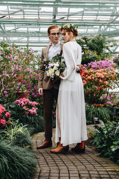 Повний вигляд красивої молодої елегантної весільної пари, що стоїть між квітами в ботанічному саду — стокове фото
