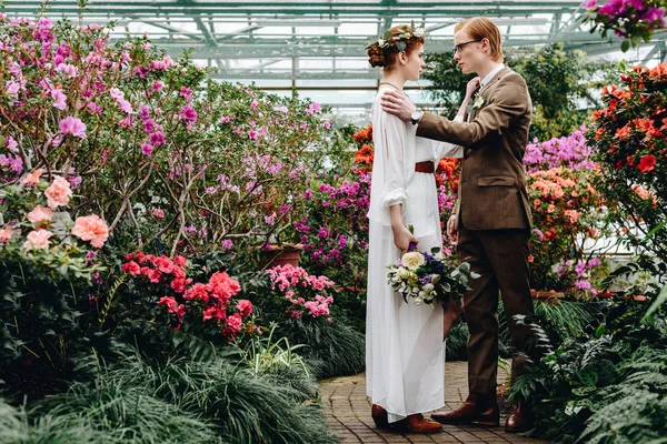 Side view of beautiful stylish young wedding couple looking at each other while standing between flowers in botanical garden — Stock Photo