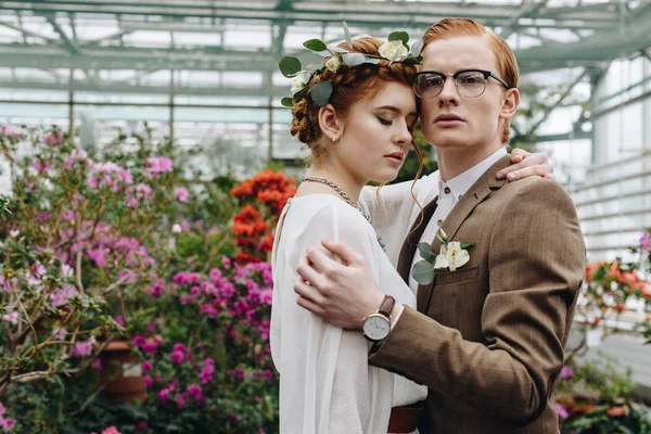 Нежная молодая рыжая свадебная пара обнимается, стоя между цветами в ботаническом саду — стоковое фото