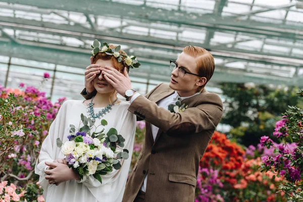 Elegante joven novio en gafas cerrando los ojos a la hermosa novia pelirroja con ramo de boda - foto de stock
