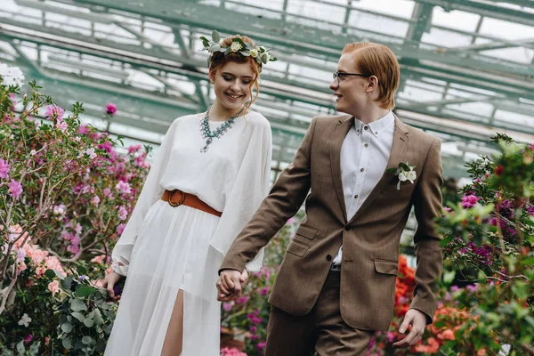 Feliz joven pelirroja boda pareja cogida de la mano y caminando entre flores en el jardín botánico - foto de stock