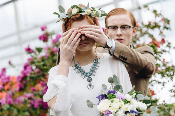 Молодой человек в очках закрывает глаза на красивую рыжую невесту со свадебным букетом — стоковое фото