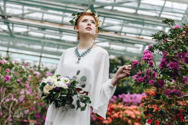 Bella giovane donna in abito bianco e corona floreale tenendo bouquet da sposa in giardino botanico — Foto stock