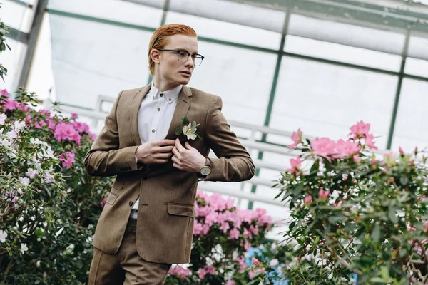 Bonito elegante jovem noivo ruiva em óculos vestindo boutonniere e olhando para longe no jardim botânico — Fotografia de Stock