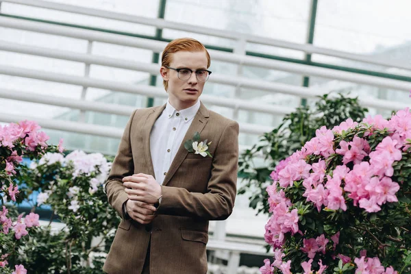 Handsome stylish young redhead groom in eyeglasses looking away in botanical garden — Stock Photo
