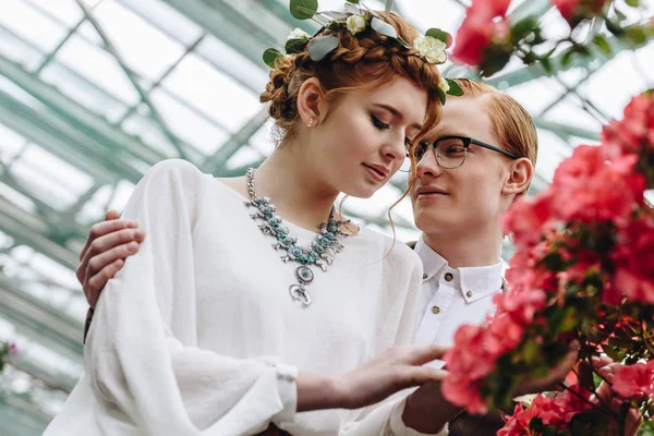 Happy young groom in eyeglasses looking at beautiful redhead bride in floral wreath — Stock Photo