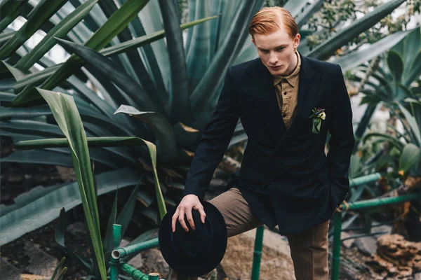 Schöner, stylischer junger rothaariger Mann mit Boutonniere, der Hut hält und im botanischen Garten steht — Stockfoto
