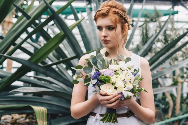 Hermosa joven pelirroja novia celebración de ramo de bodas y mirando hacia otro lado en el jardín botánico - foto de stock