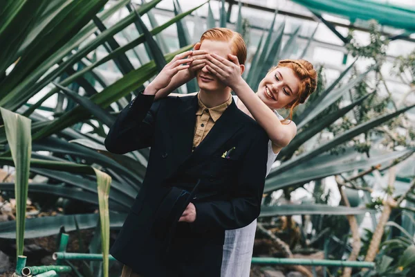 Smiling redhead girl in white dress closing eyes to young man in botanical garden — Stock Photo