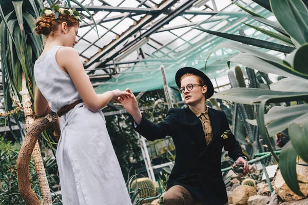Стильний молодий чоловік в капелюсі і окулярах робить пропозицію красивій дівчині в ботанічному саду — стокове фото