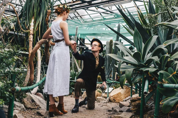 Молодий чоловік з коробкою для ювелірних виробів стоїть на коліні і тримає руку красивої молодої жінки в ботанічному саду — стокове фото