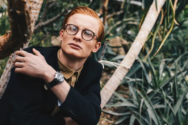 Pensive stylish young redhead man in eyeglasses looking away in botanical garden — Stock Photo