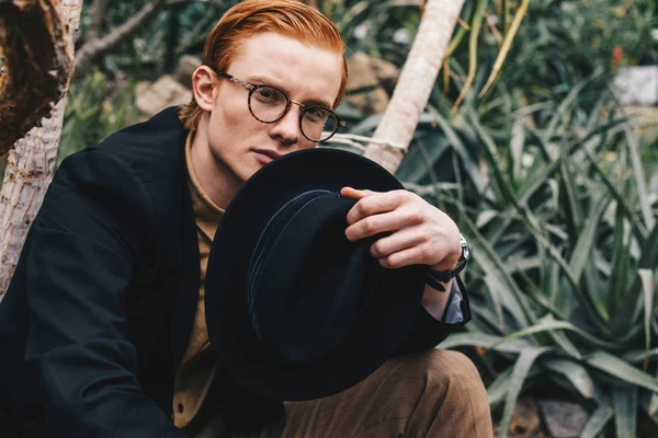 Handsome young redhead man in eyeglasses holding hat and looking at camera — Stock Photo