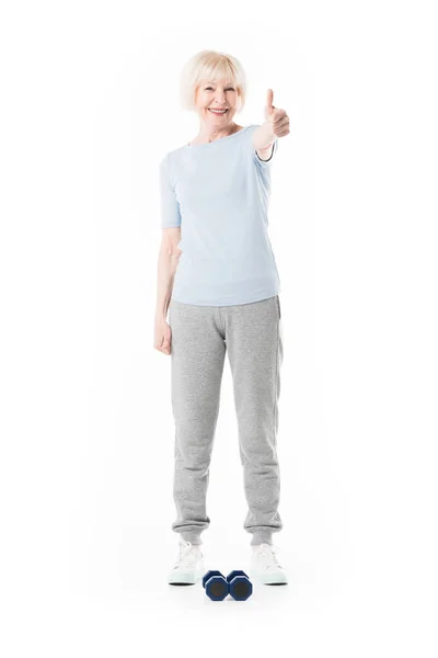 Senior sportswoman showing thumb up gesture and dumbbells on floor isolated on white — Stock Photo