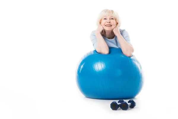 Sportiva anziana sorridente con gomiti sulla palla fitness e manubri sul pavimento isolati sul bianco — Foto stock