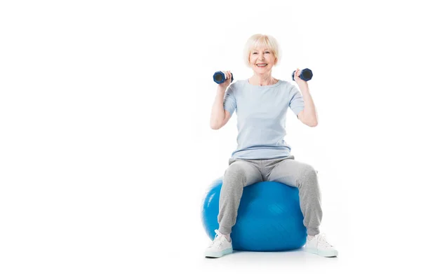 Smiling senior sportswoman with dumbbells sitting on fitness ball isolated on white — Stock Photo