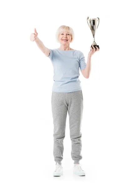 Smiling senior sportswoman showing thumb up gesture and holding trophy isolated on white — Stock Photo