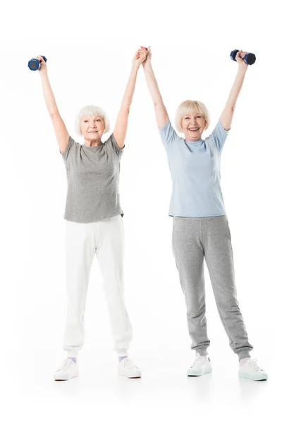 Duas desportistas seniores com braços para cima segurando halteres isolados em branco — Fotografia de Stock