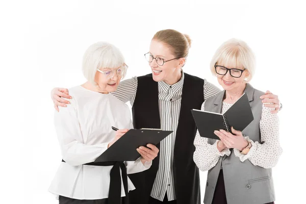 Dos mujeres de negocios con estilo senior que escriben en portapapeles y libro de texto mientras la mujer de negocios madura sostiene sus hombros aislados en blanco - foto de stock