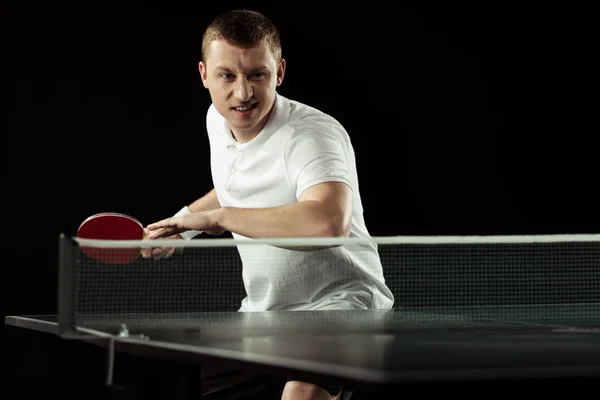 Joven jugador de tenis emocional en uniforme practicando en tenis de mesa aislado en negro - foto de stock