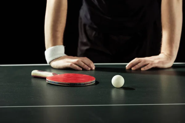 Partial view of tennis player leaning on tennis table with racket and ball isolated on black — Stock Photo