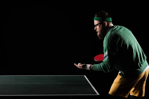 Vue latérale du joueur de tennis barbu avec raquette de tennis et balle debout à la table isolé sur noir — Photo de stock