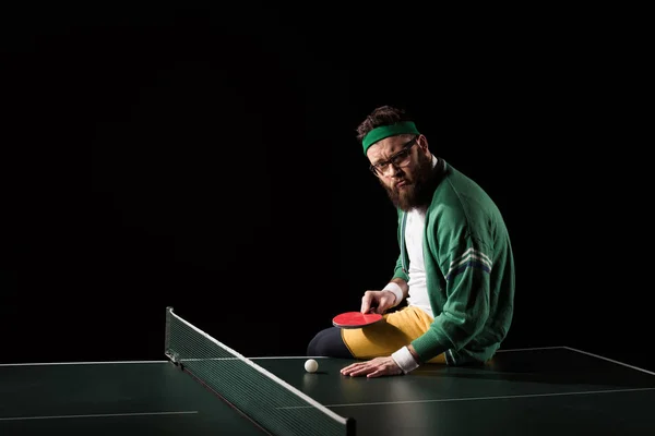 Vue latérale du joueur de tennis barbu avec raquette à la main assis sur la table isolé sur noir — Photo de stock