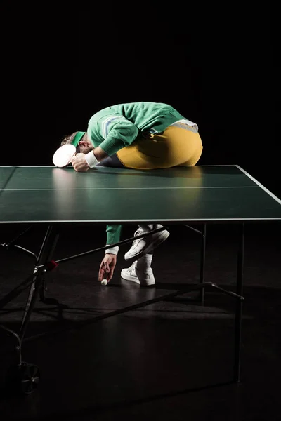 Vue arrière du joueur de tennis ramassant la balle tout en étant assis sur la table de tennis isolé sur noir — Photo de stock