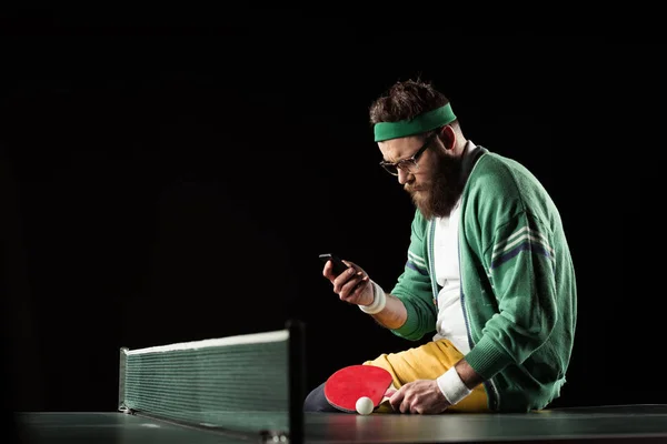 Tennis player using smartphone while sitting on tennis table isolated on black — Stock Photo