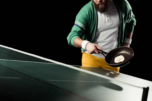 Partial view of tennis player holding frying pan with egg in hand at tennis table isolated on black — Stock Photo