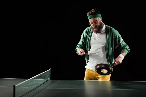 Bearded sportsman with tennis equipment and egg on frying pan isolated on black — Stock Photo
