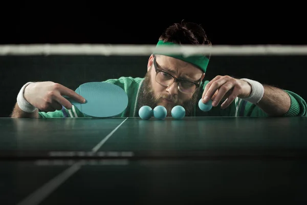 Jugador de tenis barbudo mirando pelotas de tenis azul en la mesa aislado en negro - foto de stock