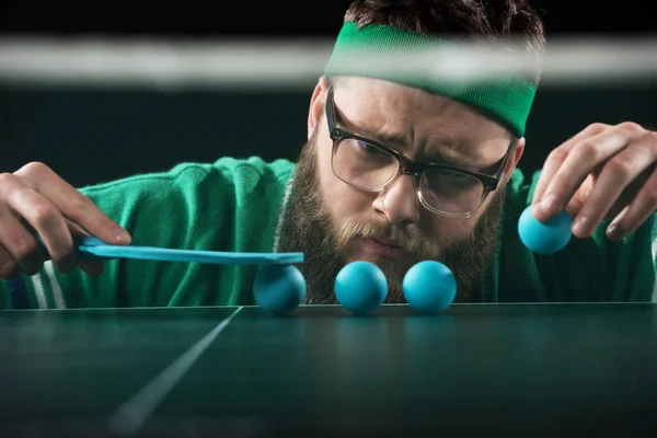 Giocatore di tennis barbuto guardando palline da tennis blu su tavolo isolato su nero — Foto stock