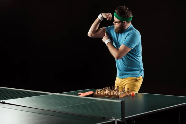 Tennis player showing muscles while standing at tennis table with chess board isolated on black — Stock Photo