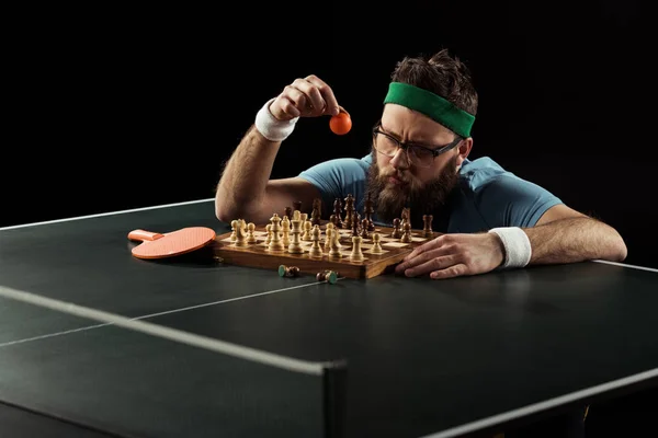 Bearded sportsman throwing tennis ball on chess board on tennis table isolated on black — Stock Photo