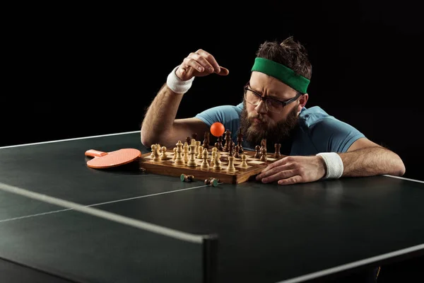 Focused bearded tennis player throwing ball on chess board on tennis table isolated on black — Stock Photo