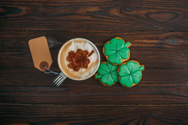 Vista superior de la taza de café con etiqueta en blanco y galletas en forma de tréboles en la mesa de madera - foto de stock