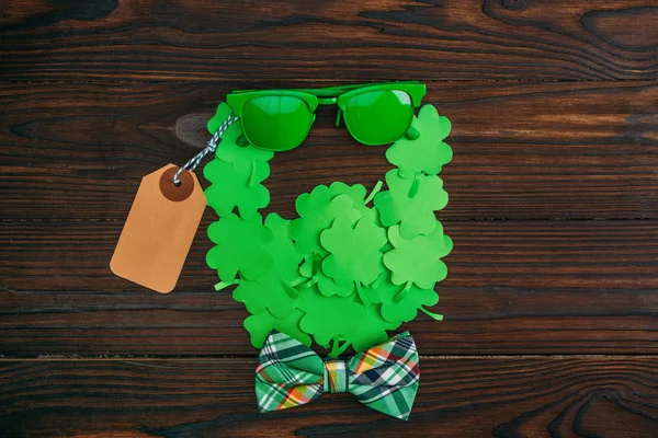 Vue du dessus du symbole du visage barbu en trèfles verts, lunettes de soleil, étiquette vierge et noeud papillon sur une table en bois — Photo de stock