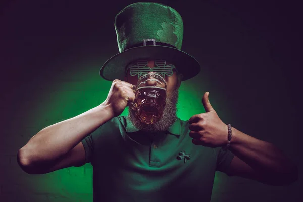Bearded man in green irish hat drinking beer and showing thumb up — Stock Photo