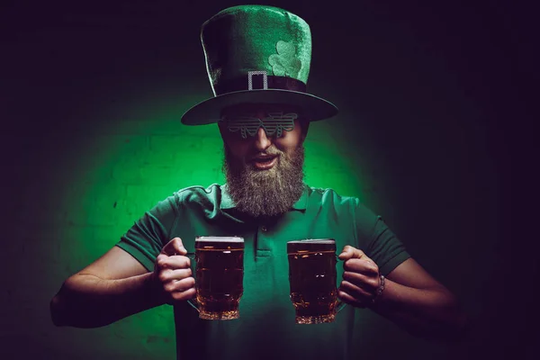 Hombre barbudo sonriente en sombrero irlandés verde sosteniendo vasos de cerveza - foto de stock