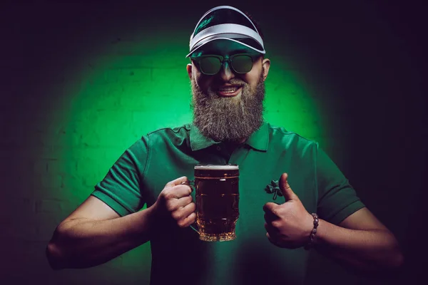 Sonriente barbudo hombre sosteniendo vaso de cerveza y mostrando el pulgar hacia arriba, st patricks concepto del día - foto de stock